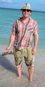 person standing ankle deep in miami beach ocean short sleeve plaid shirt knee length olive green shorts holding sandles wearing fedora